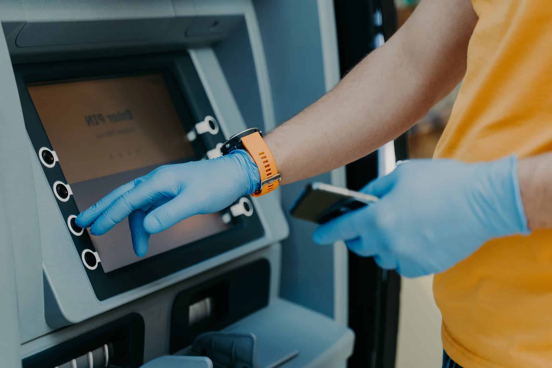 Cropped shot of man wears protective rubber gloves, uses ATM machie to withdraw money, prevents from corona virus or covid-19, holds mobile phone, presses button. Virus pandemic. Health care concept