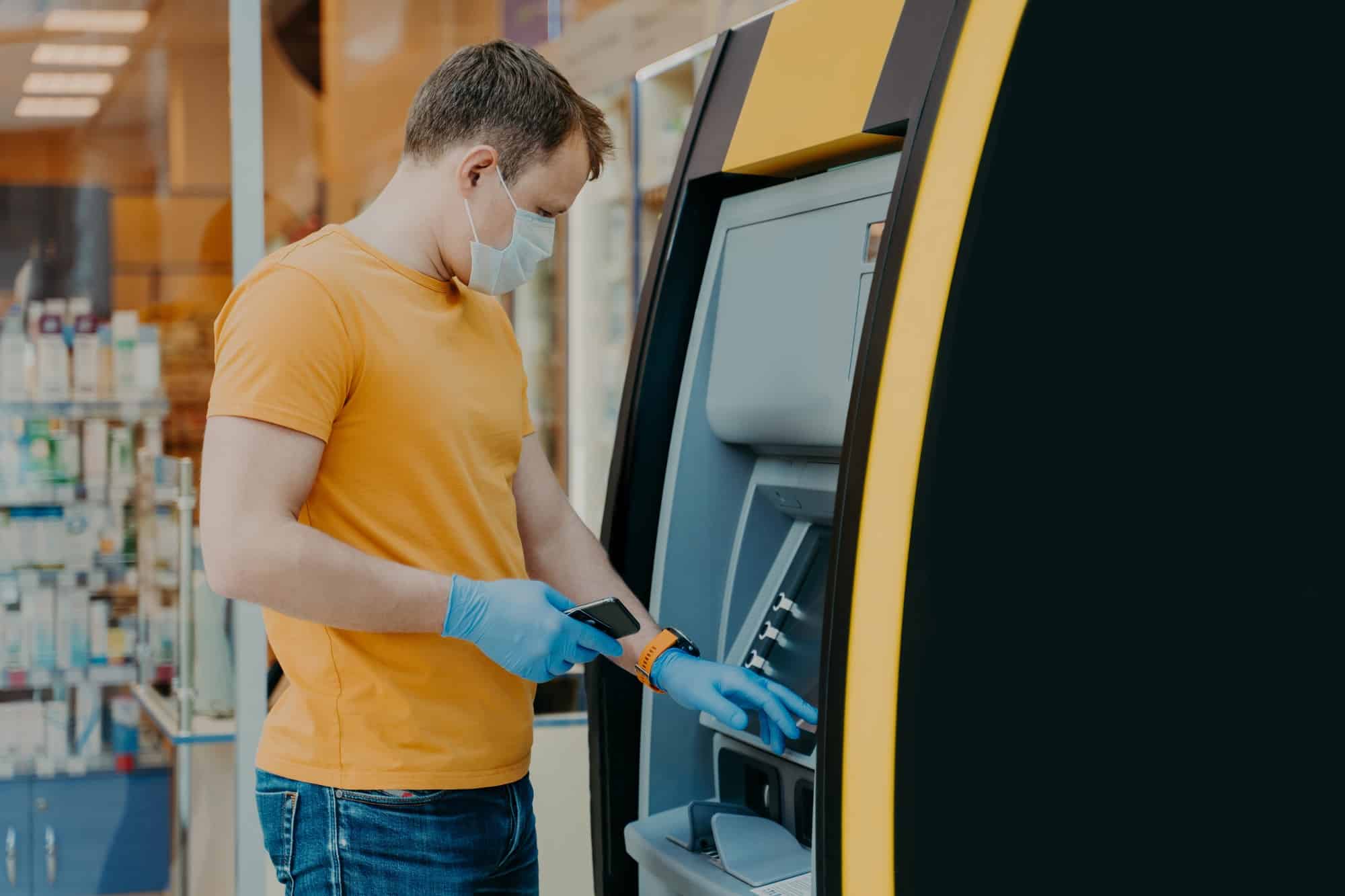 Man wears protective mask and medical gloves, uses ATM machine, prevents from virus pandemic